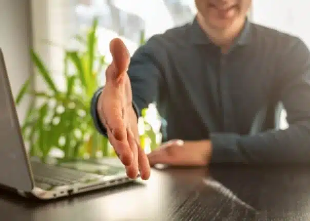 Eine Person bietet einen Handschlag an, daneben steht ein Laptop.