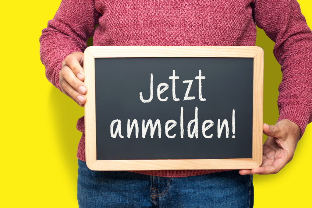 Person in rotem Pullover steht vor gelbem Hintergrund und hält eine kleine Tafel mit der Aufschrift „Jetzt anmelden!“.