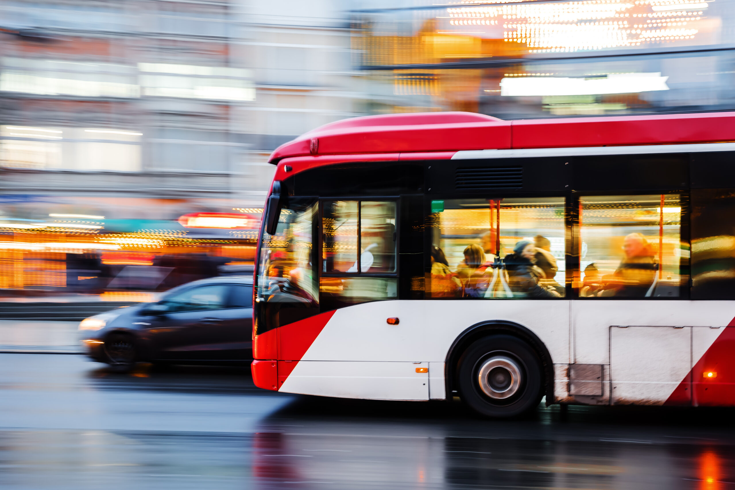 Ein rot-weißer Stadtbus fährt auf nasser Straße, im Hintergrund sieht man verschwommene Lichter der Stadt.
